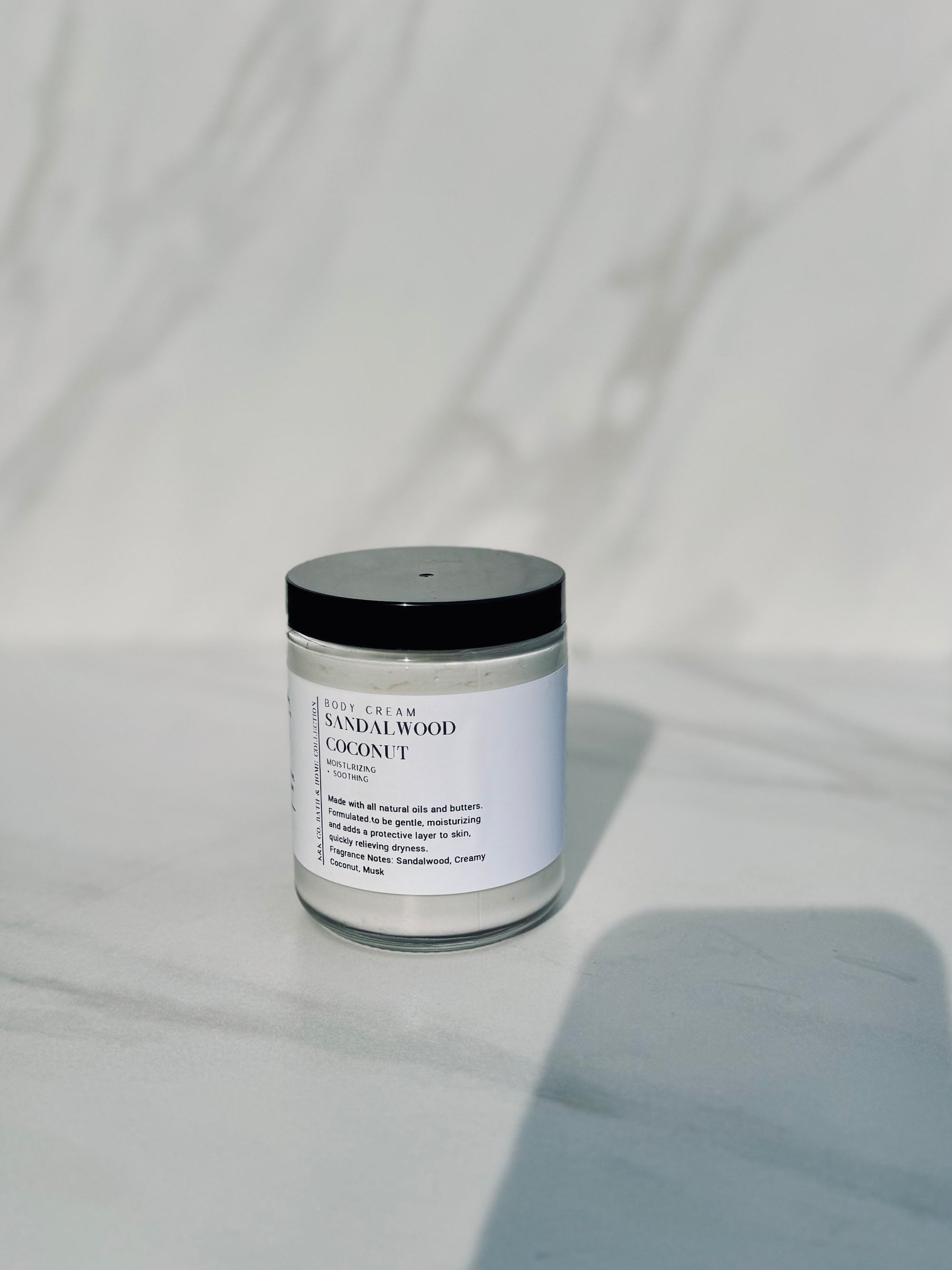 A jar of Sandalwood Coconut Body Cream placed on a marble surface with shadows falling over the frame.
